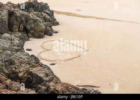 Cuore su una spiaggia di sabbia con rock come sfondo in una composizione romantica Foto Stock