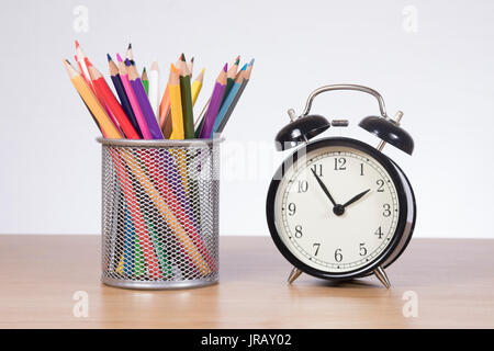 Cestello in filo metallico con matite colorate e un vecchio stile della campana di allarme orologio su una scrivania di legno in una immagine concettuale Foto Stock