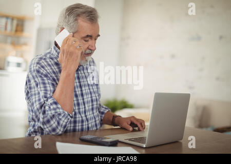 Preoccupato senior man tenendo il telefono mentre utilizzando portatile a casa Foto Stock