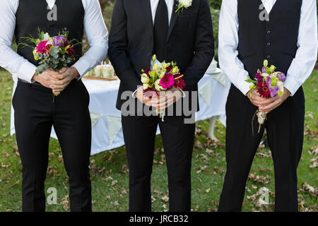 Sezione intermedia dello sposo e della miglior uomo in piedi con bouquet di fiori nel giardino Foto Stock