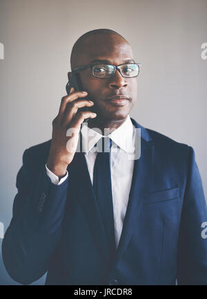 Ritratto di un fiducioso giovane imprenditore africano indossa una tuta e occhiali parlando al cellulare mentre sta in piedi da solo in un ufficio Foto Stock