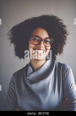 Ritratto di un sorridente giovane imprenditrice africana con gli occhiali e stare in piedi da sola iin un ufficio con le braccia incrociate Foto Stock