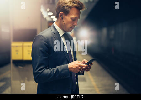 Giovane imprenditore che indossa gli auricolari e la lettura dei messaggi di testo sul suo cellulare mentre in piedi su una piattaforma della metropolitana durante i suoi spostamenti di mattina Foto Stock