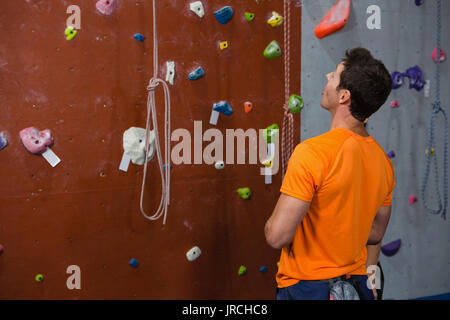 Vista posteriore del giovane uomo che guarda verso l'alto mentre in piedi dalla parete di arrampicata in palestra Foto Stock