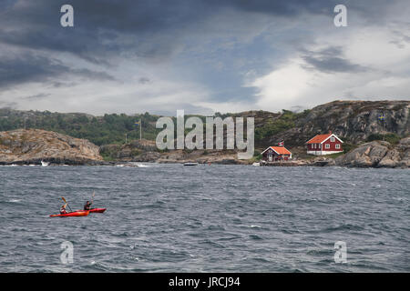 Due persone in kayak in mare nella costa della Svezia con le tipiche case rosse e piccole colline di roccia si vede in background con il buio e la luce cloud Foto Stock