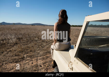 Vista posteriore della donna seduta sul cofano del veicolo mentre guarda il paesaggio Foto Stock