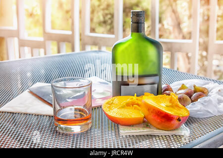 Mango e figure con una bottiglia e bicchiere di brandy sul tavolo sul balcone in una giornata di sole. Il telaio orizzontale. Foto Stock