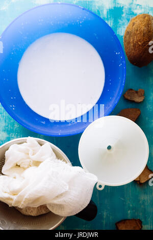 Vista dall'alto di un n in casa di latte di cocco mescolato in un frullatore e pressato attraverso un setaccio di cotone, versata in una ciotola blu , con utensili da cucina e una noce di cocco reale Foto Stock