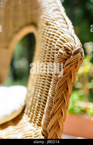 Vista ravvicinata sul bracciolo di una poltrona in rattan con cuscino bianco alla luce del sole su un balcone con piantatrice. Foto Stock