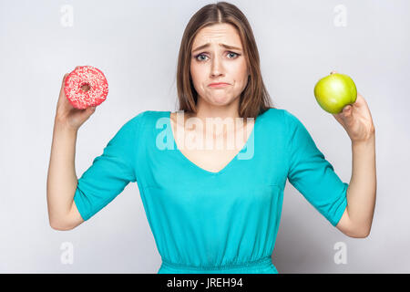 Bella giovane donna con lentiggini in abito verde, confussed e cercando di fare una scelta tra apple e ciambella. studio shot su sfondo grigio chiaro Foto Stock