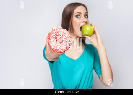 Bella giovane donna con lentiggini in abito verde, mangiare mela verde e rosa di contenimento ciambella. studio shot su sfondo grigio chiaro Foto Stock