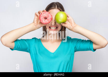 Bella giovane donna con lentiggini in abito verde, tenendo davanti agli occhi della mela verde e rosa donut e mostra i suoi denti. studio shot su grigio chiaro Foto Stock