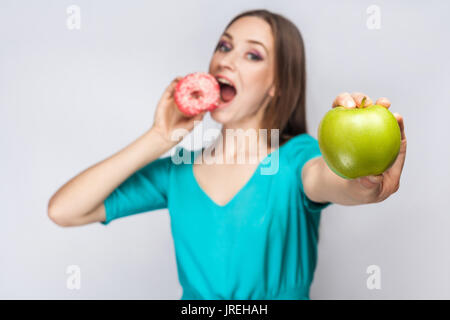 Bella giovane donna con lentiggini in abito verde, mangiare ciambella rosa e tenendo premuto mela verde. studio shot su sfondo grigio chiaro Foto Stock