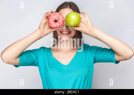 Bella giovane donna con lentiggini in abito verde, tenendo davanti agli occhi della mela verde e rosa ciambella con sorriso toothy. studio shot su grigio chiaro ba Foto Stock