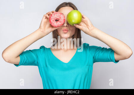 Bella giovane donna con lentiggini in abito verde, tenendo davanti agli occhi della mela verde e rosa a ciambella e baciare. studio shot su grigio chiaro backgrou Foto Stock