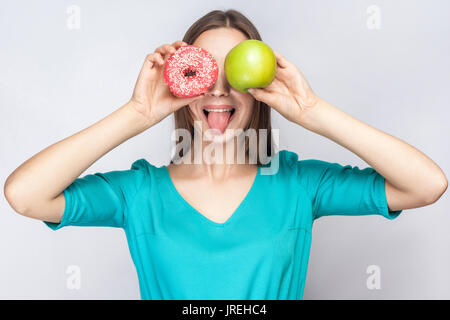 Bella giovane donna con lentiggini in abito verde, tenendo davanti agli occhi della mela verde e rosa donut e mostra la linguetta. studio shot su grigio chiaro bac Foto Stock