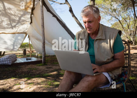 Uomo maturo utilizzando computer portatile mentre è seduto su una sedia al campeggio Foto Stock