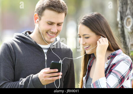 Felice coppia di adolescenti la condivisione on line di musica e la visione di contenuti multimediali in uno smart phone all'aperto in un parco Foto Stock