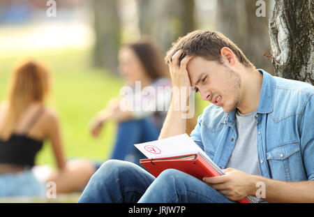 Singolo studente triste la verifica di un esame non superato seduto sull'erba in un parco con persone non focalizzato in background Foto Stock