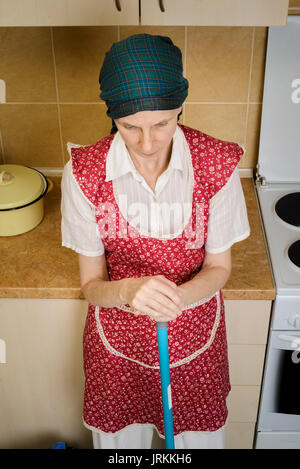 Premuto una donna adulta, una casalinga o una cameriera, indossare un grembiule rosso e un foulard verde sul suo capo è a riposo dopo che ella ha spazzato la cucina con una b Foto Stock