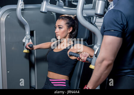 Sport e fitness, il lavoro di squadra e il concetto di persone - una giovane donna muscoli di flessione sulla macchina di palestra e personal trainer con clipboard Foto Stock