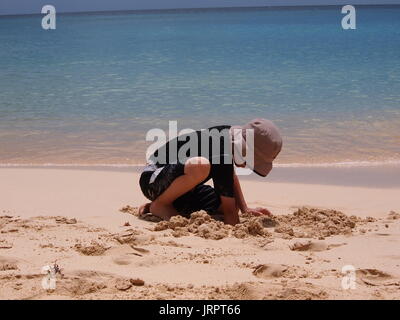 Ragazzo che indossa un cappello e top nero di scavare un buco in spiaggia, Meads Bay, Anguilla BWI. Foto Stock