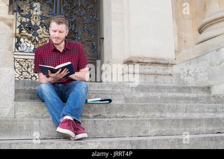 Università maschio studente seduto sui gradini e la lettura fuori dall'edificio Foto Stock