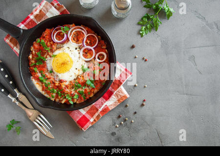 Shakshuka in una ghisa Pan. Stile medio orientale le uova in camicia in salsa di pomodoro e peperone con prezzemolo e cipolla e spezie sul cemento grigio Sfondo. Foto Stock