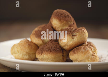 Closeup colpo di profiteroles ricoperti di polvere di cacao su piastra bianca sul tavolo di legno Foto Stock