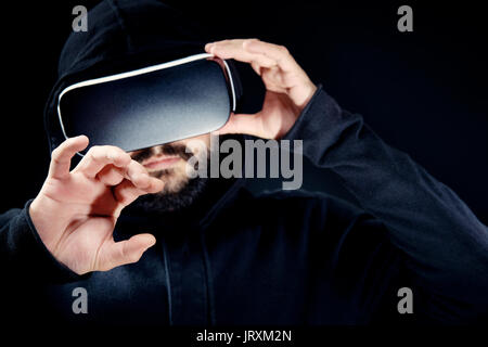 Giovani hipster barbuto man in black top con cappuccio, indossando la realtà virtuale gli occhiali. Sfondo nero studio concetto VR Foto Stock