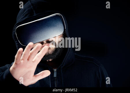 Giovani hipster barbuto man in black top con cappuccio, indossando la realtà virtuale gli occhiali. Sfondo nero studio concetto VR Foto Stock
