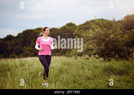 Montare la donna di mezza età a piedi attraverso un campo rurale ascolta musica sul suo telefono mobile utilizzando gli auricolari guardando al lato con un sorriso in un h Foto Stock