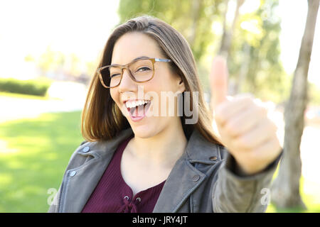 Ritratto di un funny girl indossando occhiali gesticolando Thumbs up e guardando a voi all'aperto in un parco Foto Stock