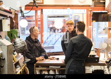 Cobbler che ripara scarpe in pelle all'interno di un'officina tradizionale. Sulla parete sono visibili utensili, attrezzature di taglio chiave e suole di ricambio Foto Stock