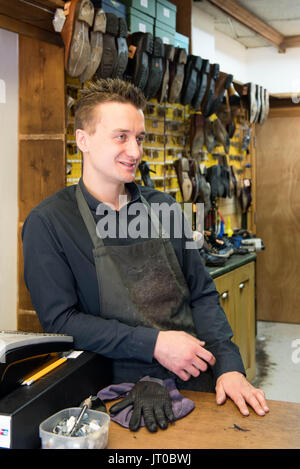 Cobbler che ripara scarpe in pelle all'interno di un'officina tradizionale. Sulla parete sono visibili utensili, attrezzature di taglio chiave e suole di ricambio Foto Stock