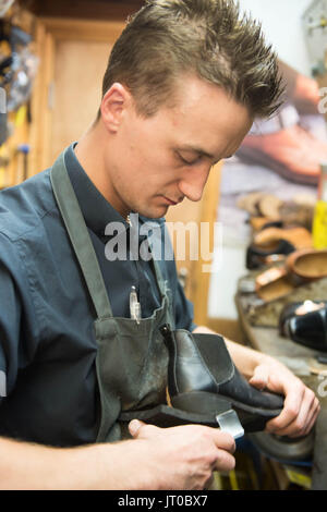 Cobbler che ripara scarpe in pelle all'interno di un'officina tradizionale. Sulla parete sono visibili utensili, attrezzature di taglio chiave e suole di ricambio Foto Stock