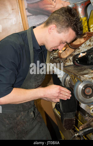Cobbler che ripara scarpe in pelle all'interno di un'officina tradizionale. Sulla parete sono visibili utensili, attrezzature di taglio chiave e suole di ricambio Foto Stock