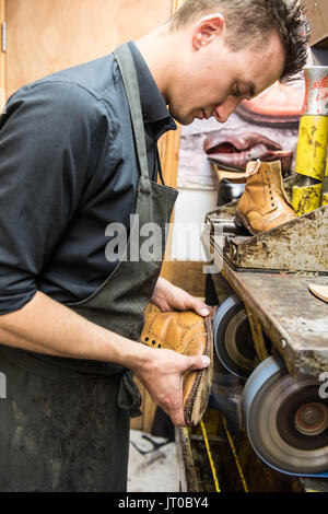 Cobbler che ripara scarpe in pelle all'interno di un'officina tradizionale. Sulla parete sono visibili utensili, attrezzature di taglio chiave e suole di ricambio Foto Stock