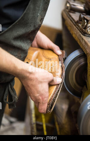 Cobbler che ripara scarpe in pelle all'interno di un'officina tradizionale. Sulla parete sono visibili utensili, attrezzature di taglio chiave e suole di ricambio Foto Stock