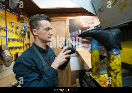 Cobbler che ripara scarpe in pelle all'interno di un'officina tradizionale. Sulla parete sono visibili utensili, attrezzature di taglio chiave e suole di ricambio Foto Stock