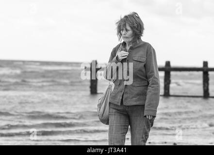 Anziani / donna di mezza età passeggiate in riva al mare sulla costa in una giornata di vento nel Regno Unito. In bianco e nero dell'immagine. Foto Stock