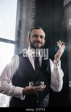 Ritratto di bello sorridente uomo barbuto azienda posacenere e accendisigari fumare in luoghi chiusi Foto Stock