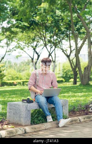 Giovane uomo seduto sulla panchina nel parco, libro, sorridente e guardando nella telecamera Foto Stock