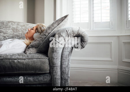 Donna matura giacente sul suo divano, godendo di ascolto di musica attraverso le cuffie. Foto Stock