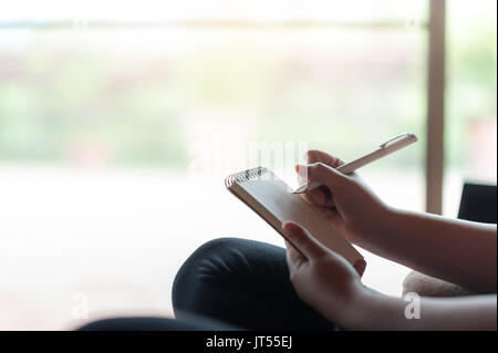 La donna la scrittura a mano qualche idea sul piccolo blocco note mentre è seduto su una sedia in casa di lavoro freelance dal concetto ovunque Foto Stock