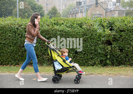 Occupata la mamma di parlare al telefono mentre si spinge il suo bambino seduto in una carrozzina. Foto Stock