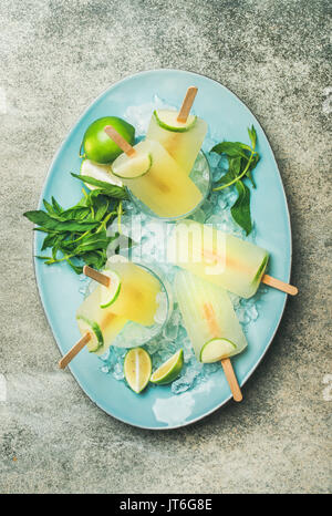 Estate rinfrescante limonata popsicles con lime e menta in bicchieri con scaglie di ghiaccio su piastra blu su sfondo grigio, vista dall'alto Foto Stock