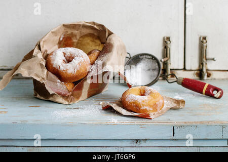 Ciambelle fresche con lo zucchero in polvere dal sacchetto di carta servita con setaccio vintage blu sul tavolo di legno. In stile rustico, la luce del giorno. Foto Stock