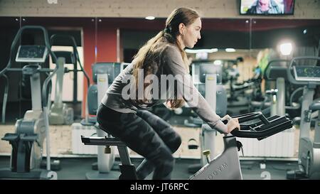 Giovane bella ragazza in palestra, agitando le gambe su Cycling Simulator, sorridente alla fotocamera. Il concetto: per amore dello sport, a frequentare una palestra, nutrizione adeguata, un corpo snello, di essere sano. 4k Foto Stock