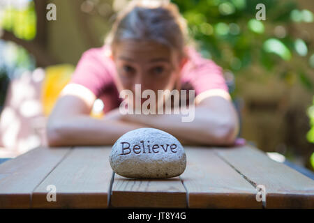 Giovane donna seduta a un tavolo di legno in background con una roccia di fronte a lei con incisa la parola credere .modello di rilascio Foto Stock